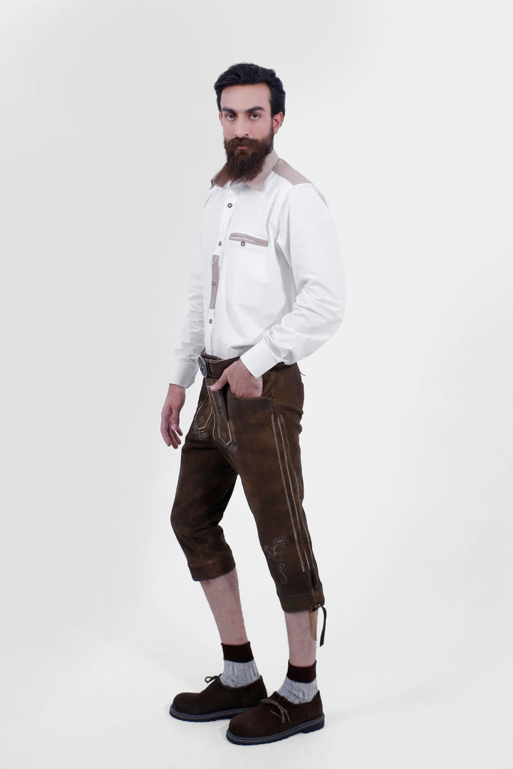 A male shown in a side profile wearing a white shirt and Oslo Bundhosen. One of his hands is in the pocket of the pants, highlighting the side embroidery and detailing, including a small side pocket. The model is wearing brown shoes with grey socks, and the mid-length pants are secured with a decorative belt.