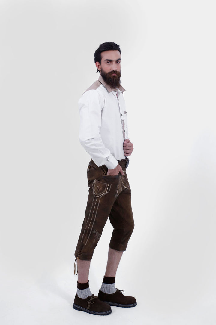 A male standing in a semi-profile pose, wearing a white shirt and Oslo Bundhosen. One hand is in his pocket, showcasing the side embroidery, small pocket details, and overall fit of the pants. With traditional Bavarian embroidery and a decorative belt. He is wearing brown shoes and grey socks