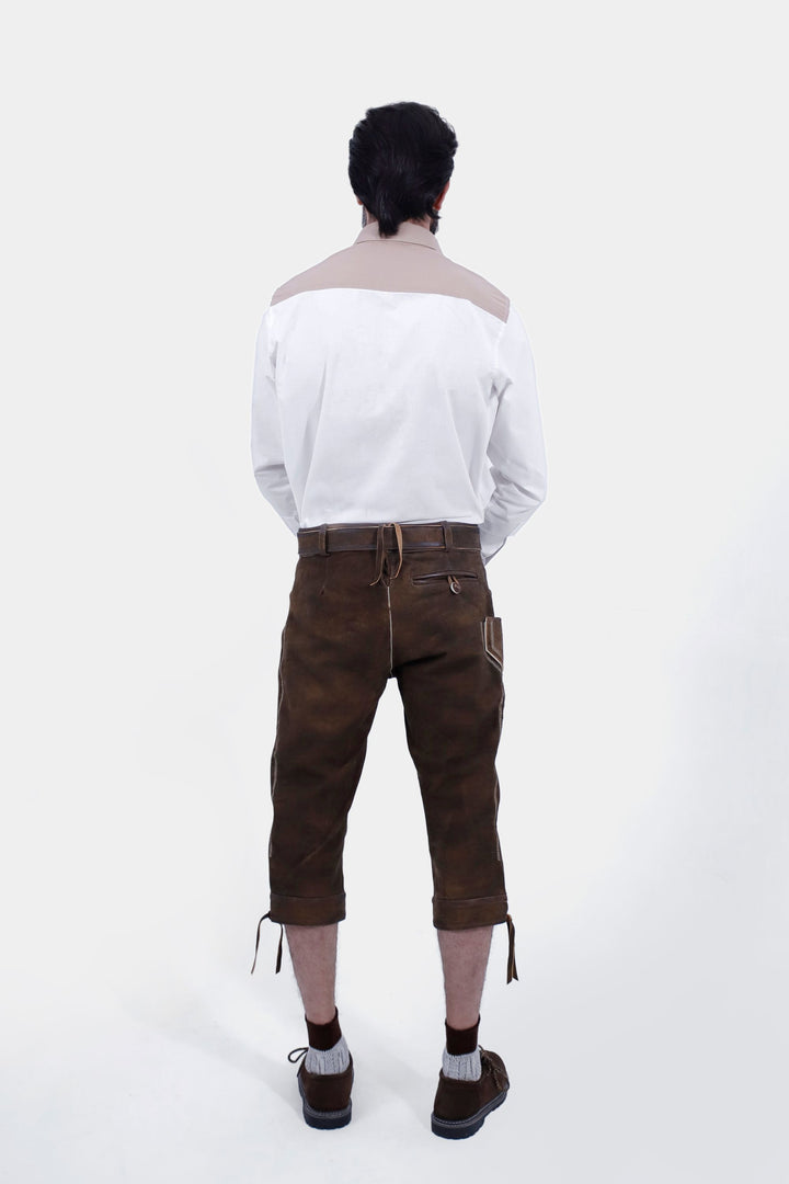 The back view of a male with a beard standing, wearing a white shirt and Oslo Bundhosen. The image focuses on the back details of the pants, including the back pockets, embroidery, and an adjustable tie at the waist. The man is also wearing brown shoes with grey socks.