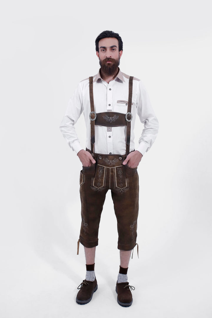   A man with a beard is wearing a white shirt and Oslo Bundhosen with suspenders. The suspenders have decorative buckles and are attached to the pants, which feature intricate front embroidery. The man stands with both hands in his pockets, wearing brown shoes and grey socks.