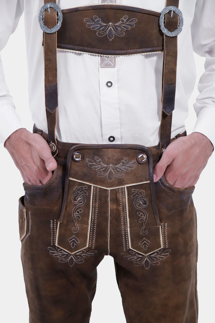 A close-up view of the front of the Oslo Bundhosen, worn by a man. The focus is on the intricate embroidery on the front of the pants, the detailed buttons on the suspenders, and the decorative belt buckle. The leather-like material and traditional Bavarian styling are clearly visible.