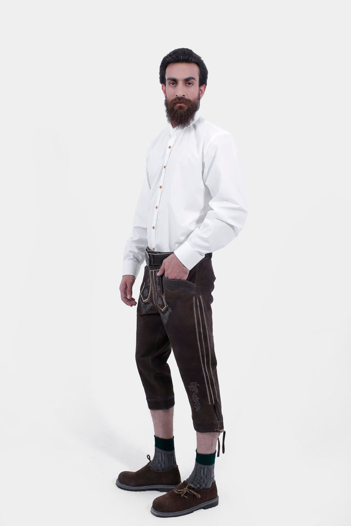 A side profile of a man showcasing Philipp Lahm Bundhosen Outfit paired with a white button-down shirt. The Bundhosen, made of dark brown leather, feature intricate embroidery and a decorative belt buckle. Paring with traditional socks and brown shoes