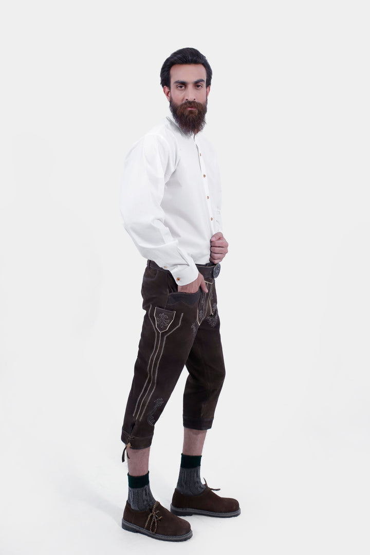 A side profile of a man showcasing Philipp Lahm Bundhosen Outfit paired with a white button-down shirt. The Bundhosen, made of dark brown leather, feature intricate embroidery and a decorative belt buckle. Paring with traditional socks and brown shoes