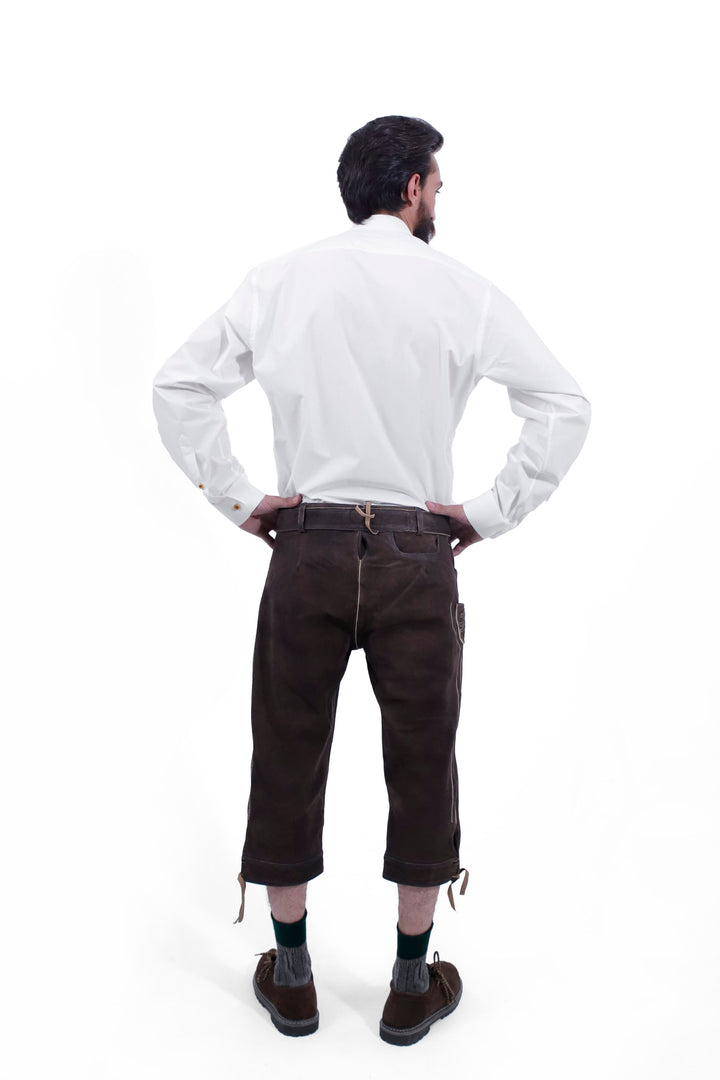 The back view of a man wearing Philipp Lahm Bundhosen Outfit and a white shirt. The craftsmanship of the dark brown leather pants is highlighted, showing the intricate details and secure fit from behind. Paring with traditional socks and brown shoes