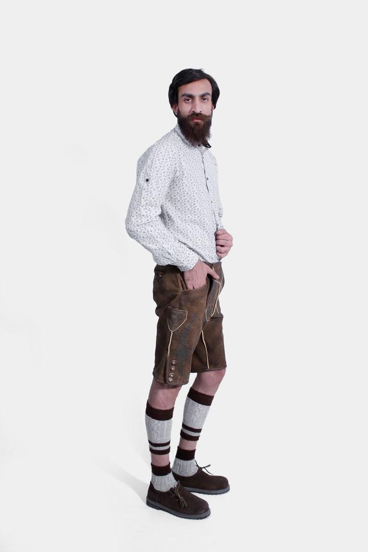 A bearded man wearing a white patterned shirt and Schubert Lederhosen, posing with his hand in his pocket. The lederhosen features detailed embroidery and button accents, paired with traditional striped knee-high socks and brown shoes.