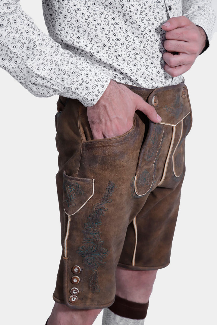 Side view of brown leather lederhosen with intricate embroidery and button details, worn by a man against a white background.