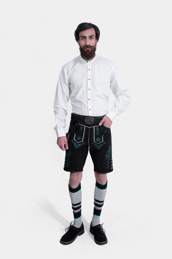 Man wearing a Schümann Lederhosen Outfit, featuring a white long-sleeved shirt with button details, black leather lederhosen with intricate green embroidery, traditional knee-high socks, and black shoes. oktoberfest outfit herren​