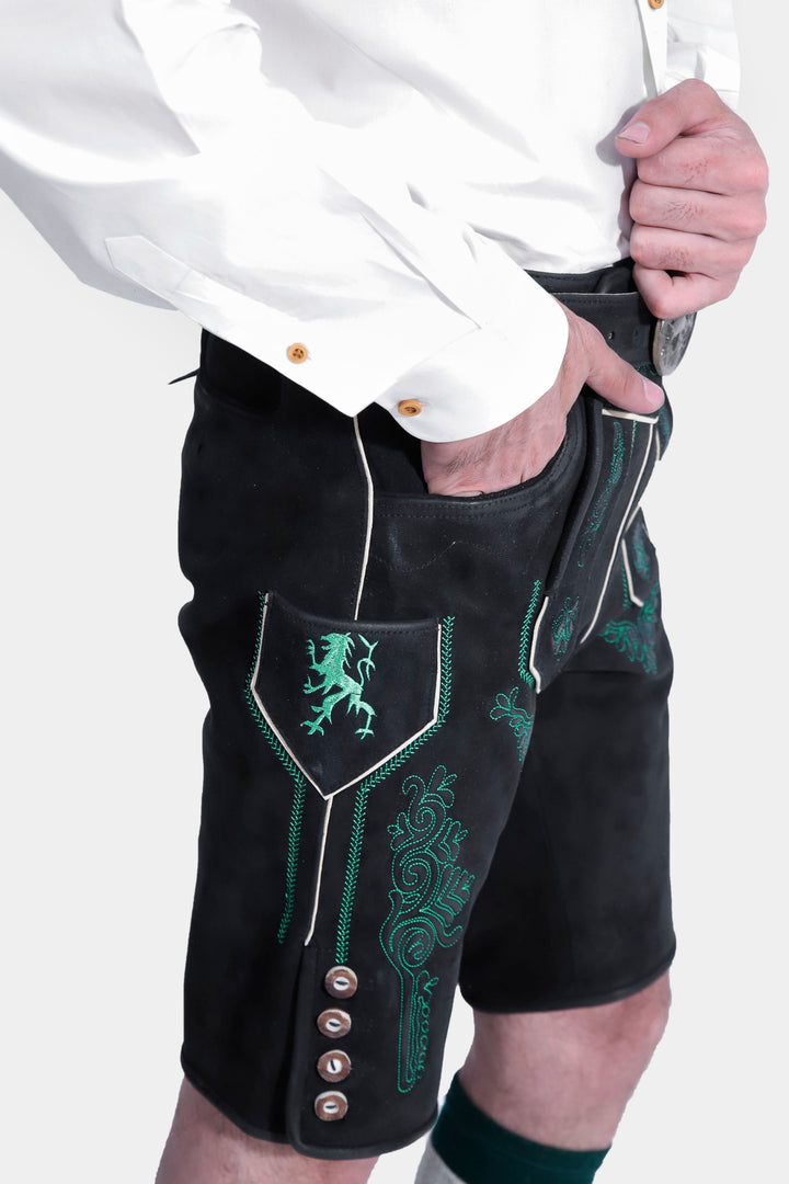 Detail shot of the Schümann Lederhosen Outfit's green embroidered pocket on the black leather lederhosen, along with the white shirt sleeve and hand resting in the pocket.