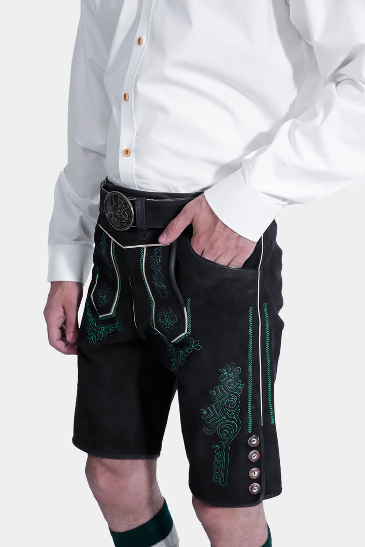 Side view of the Schümann Lederhosen Outfit, focusing on the detailed green embroidery on the black leather lederhosen, paired with a white shirt and hand in the pocket.