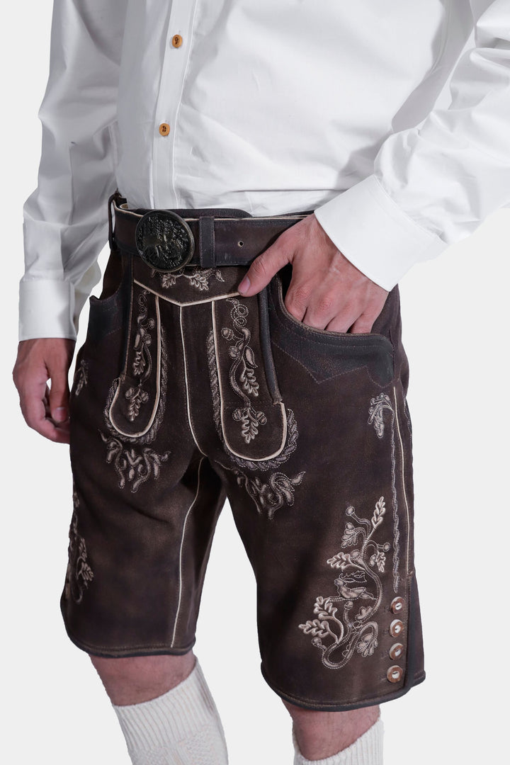 Side view of a model wearing Spiegel Lederhosen, emphasizing the detailed embroidery on the side of the leather shorts and the elegant belt buckle, paired with a white shirt.