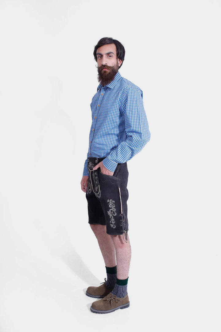 Side profile of a man in Versailles Lederhosen, emphasizing the brown leather shorts' design, embroidery, and fit, paired with a blue checkered shirt.