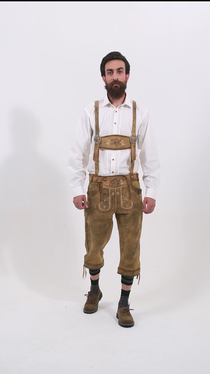 Male model wearing a white shirt and Edinburgh Bundhosen, featuring intricate embroidery on the front, a decorative belt buckle, and mid-length. The model's hands are by his sides, and he is wearing brown shoes with grey socks, showcasing the traditional bundhosen in a rotating video to display all angles.