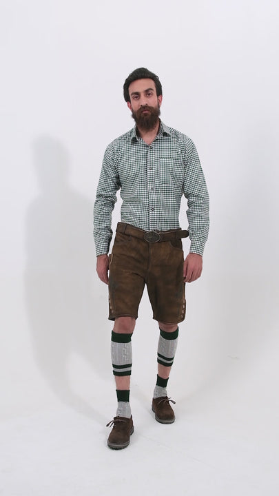 A video showcasing the Mahler Lederhosen outfit from every angle. The model, a man with a beard, wears brown leather lederhosen paired with a green checkered shirt. The video captures detailed close-ups of the embroidery, buttons, and pockets, as well as full-body views from the front, back, and sides. The model completes the look with green and white knee-high socks and brown shoes, presenting the traditional design and craftsmanship of the Mahler Lederhosen.