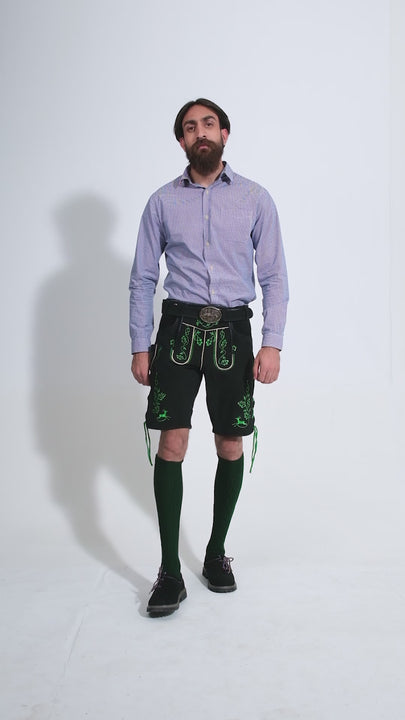 Video showcasing a man dressed in traditional black Zwinger Lederhosen with vibrant green embroidery, walking and twirling to demonstrate the outfit's fit and movement. He's paired with a purple gingham shirt and green socks, highlighting a mix of classic and modern Oktoberfest fashion.