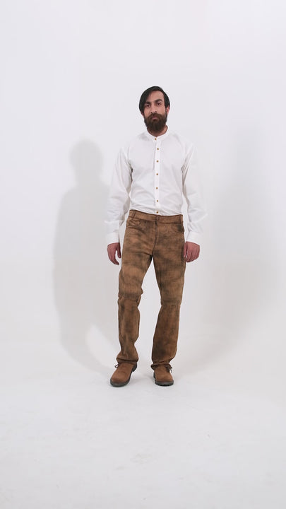 Male model wearing a white shirt and traditional brown leather bundhosen, standing with one hand in his pocket. The video showcases the bundhosen's detailed embroidery and mid-length design, highlighting the traditional craftsmanship. The model completes the look with brown shoes, presenting various angles to display the full outfit.