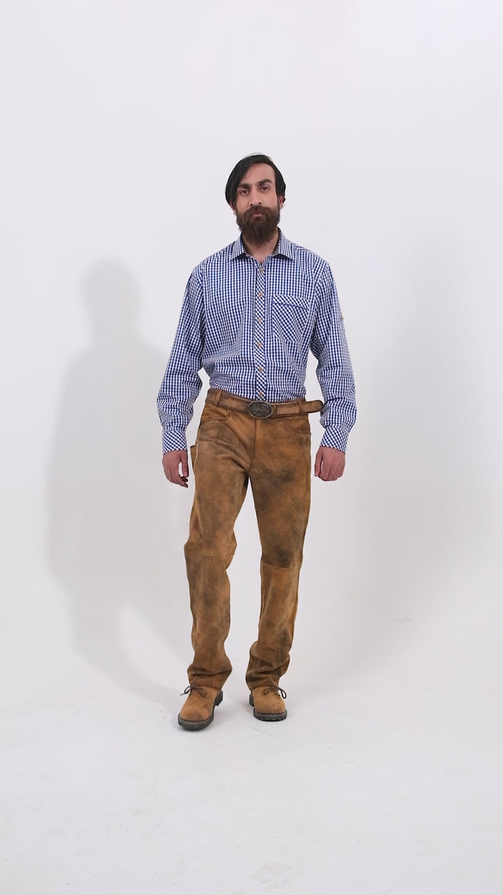 Male model wearing a blue checkered shirt and traditional brown leather bundhosen, standing with one hand in his pocket. The video highlights the bundhosen's detailed buttons and traditional design, showcasing various angles. The model completes the look with brown shoes, giving a full view of the outfit's craftsmanship.