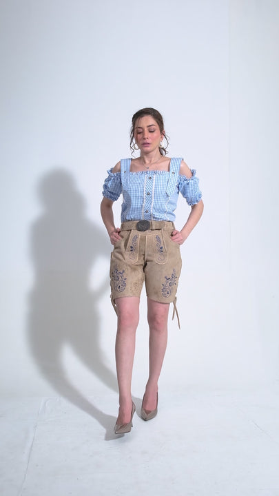 Video displaying a woman modeling beige women's Boldt Yacht Lederhosen Outfit, intricately embroidered in purple. She confidently walks and poses, showcasing the detailed craftsmanship of the embroidery and the fit of the lederhosen. The outfit is paired with a blue and white gingham off-shoulder top and sleek grey heels, combining traditional Bavarian style with a chic modern twist.