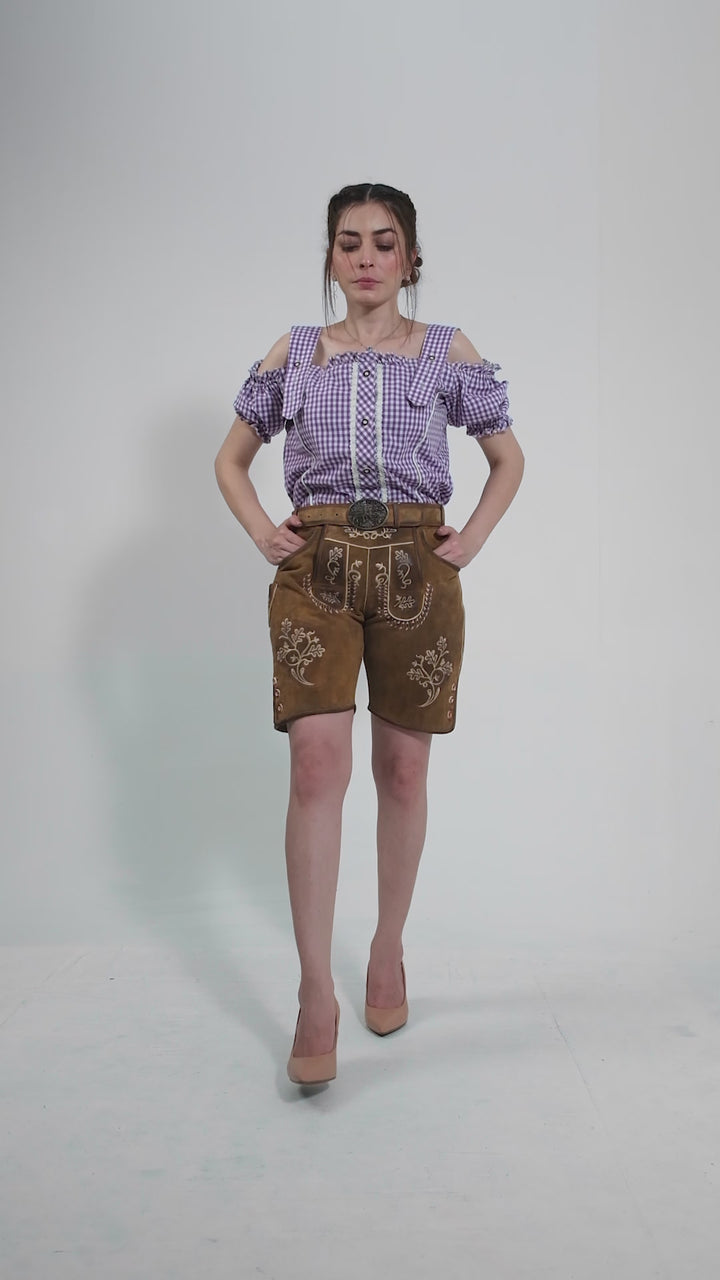 Video displaying a woman modeling traditional women's lederhosen in a taupe color, intricately embroidered with motifs. She rotates to show all angles, highlighting the detailed craftsmanship of the pockets, buttons, and embroidery. The outfit is paired with a checkered purple blouse and stylish black ankle boots, showcasing a modern twist on traditional attire.