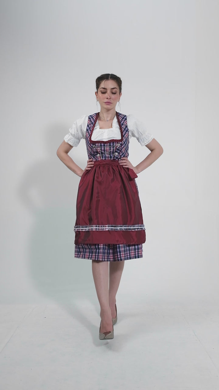 Video showcasing the Dirndl Zsa Inci, featuring a woman elegantly modeling the outfit. She highlights the vibrant crimson bodice with plaid details, the flowing skirt, and the beautifully tied apron, demonstrating the dirndl's versatility for various occasions against a neutral backdrop.