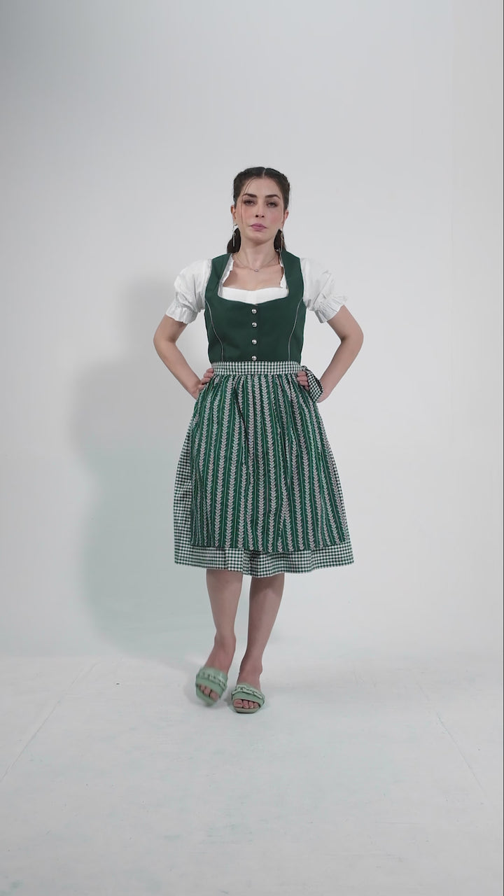 Video showcasing the Dirndl Sukowa, featuring a woman modeling the outfit with smooth movements to highlight the green bodice, patterned skirt, and checkered apron. She turns and twirls to demonstrate the flow and fit of the dirndl, set against a clean white background.