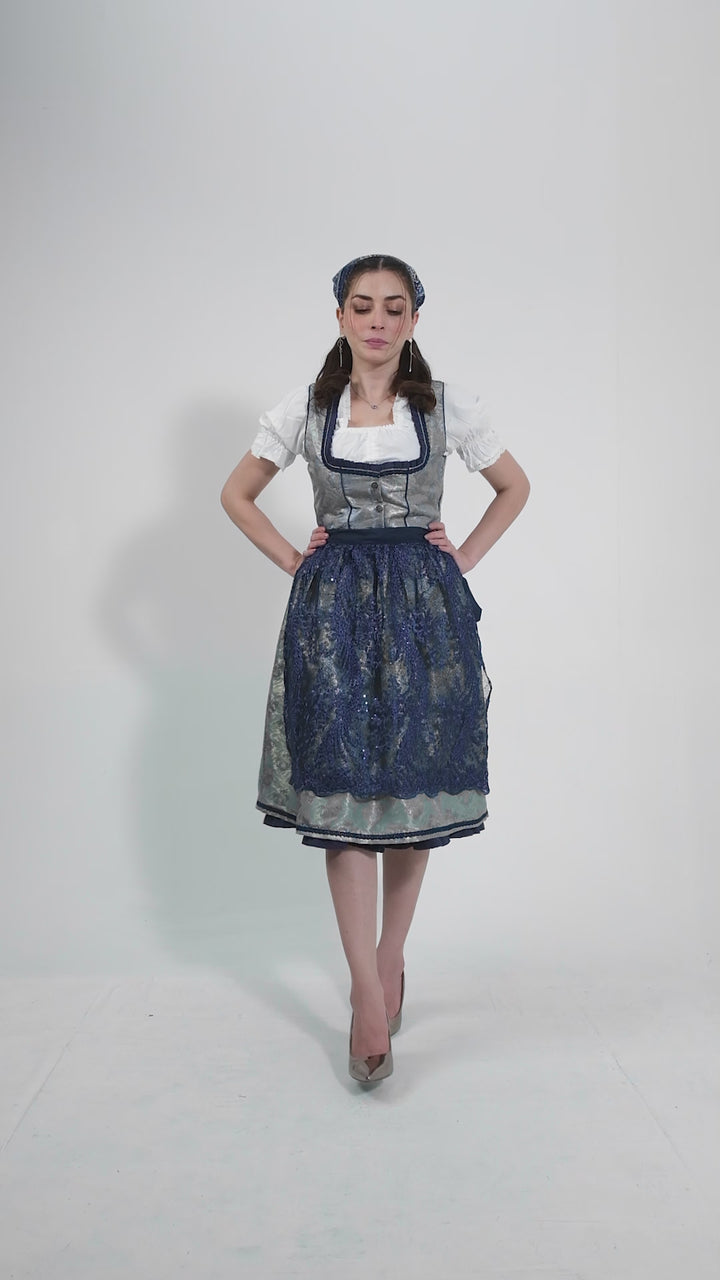 Video showcasing the Dirndl Maria Lara, featuring a woman modeling the outfit with graceful movements to highlight the intricate lace bodice, floral-patterned apron, and vibrant blue color. She twirls to demonstrate the flowing skirt, set against a neutral background.