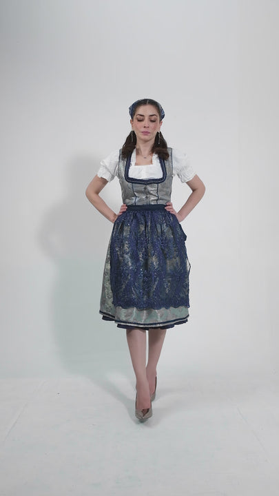 Video showcasing the Dirndl Maria Lara, featuring a woman modeling the outfit with graceful movements to highlight the intricate lace bodice, floral-patterned apron, and vibrant blue color. She twirls to demonstrate the flowing skirt, set against a neutral background.